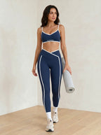 Woman in navy sports bra and leggings holding a yoga mat on a wooden floor.
