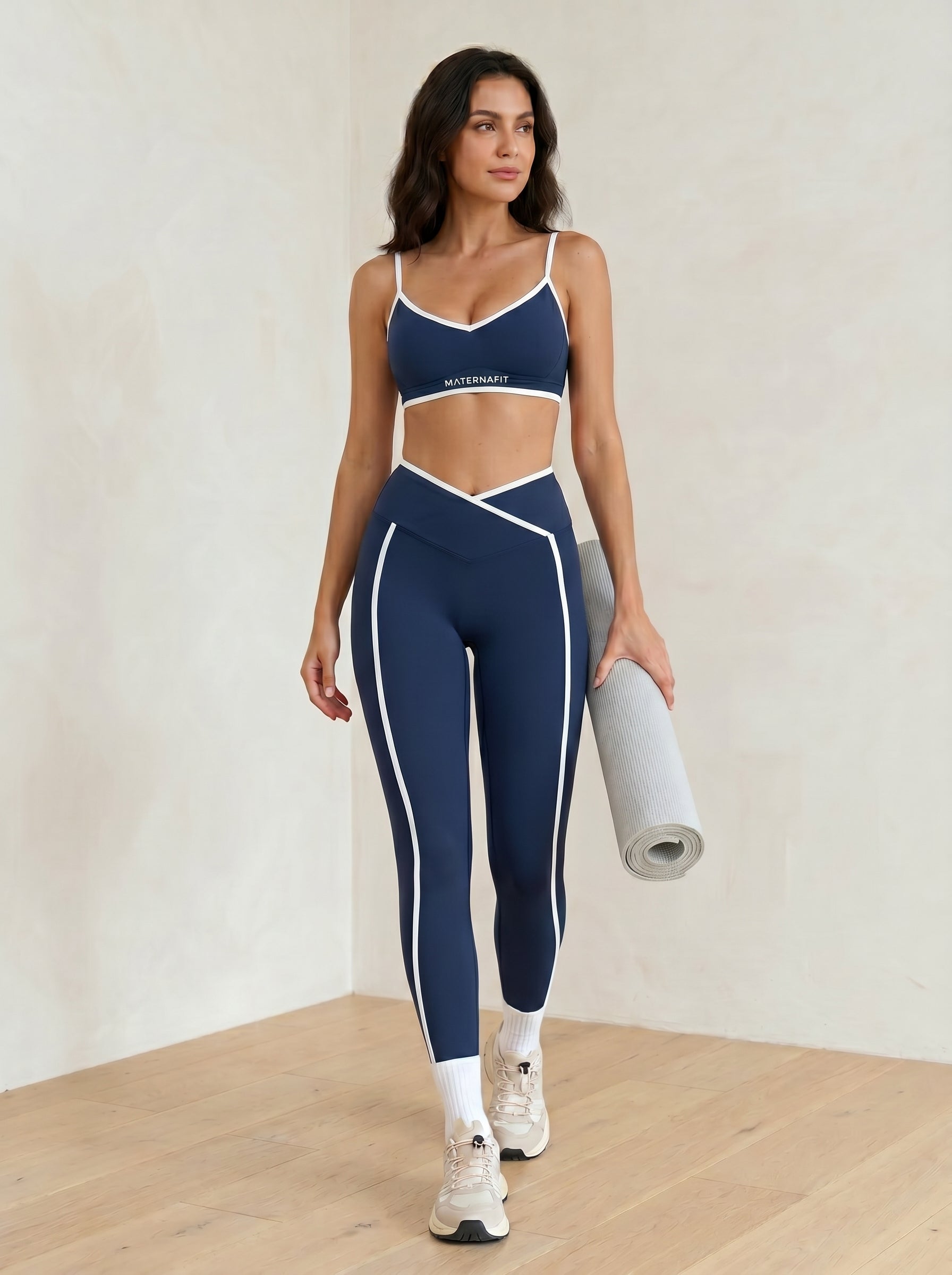Woman in navy sports bra and leggings holding a yoga mat on a wooden floor.